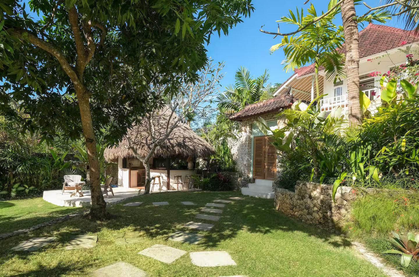 Lush green yard with stone path leading to a stone building and a thatched roof structure under a tree. Bright blue sky.