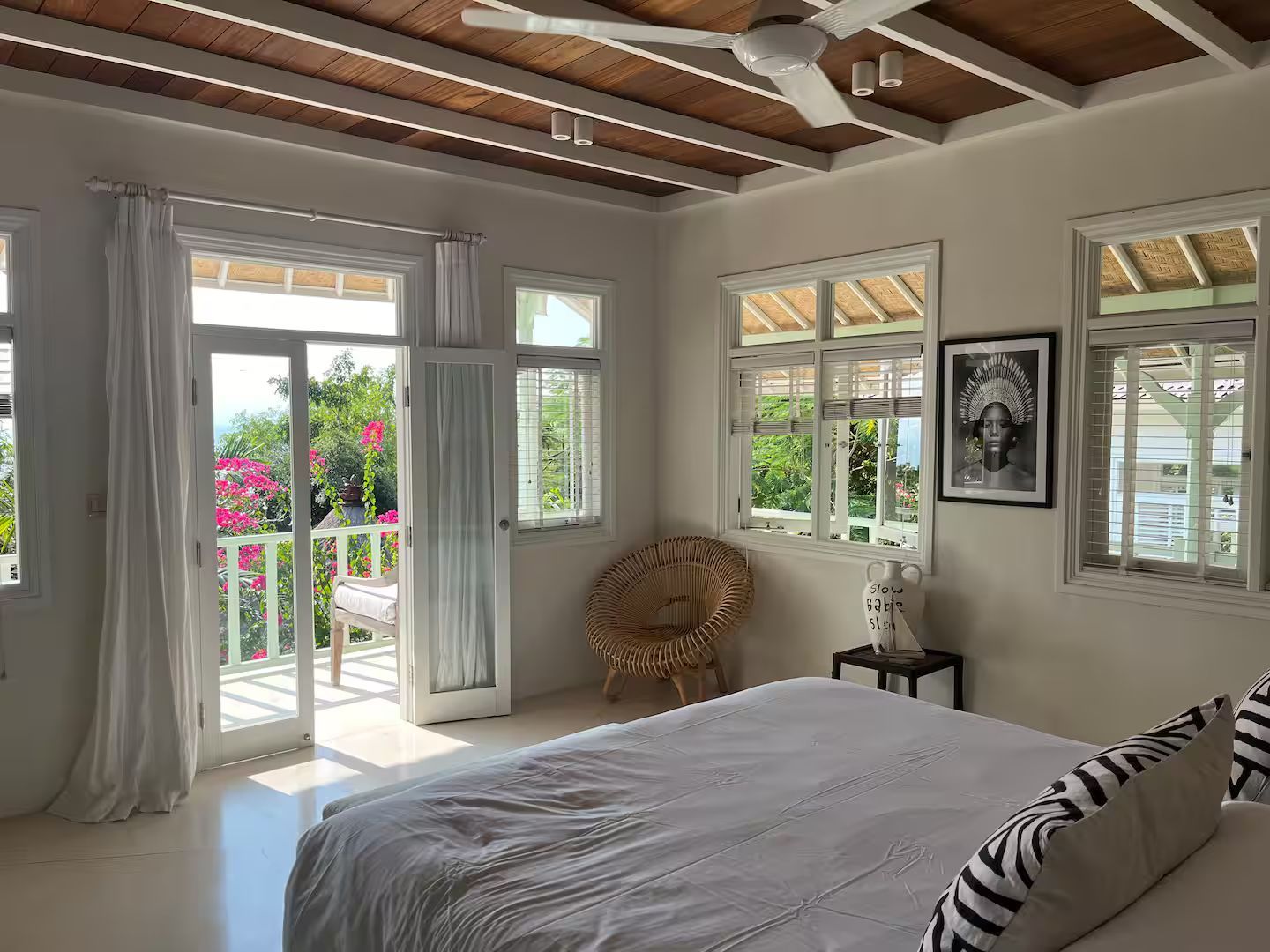 Bright bedroom with a white bed, wicker chair, and balcony overlooking greenery. Natural light fills the space.