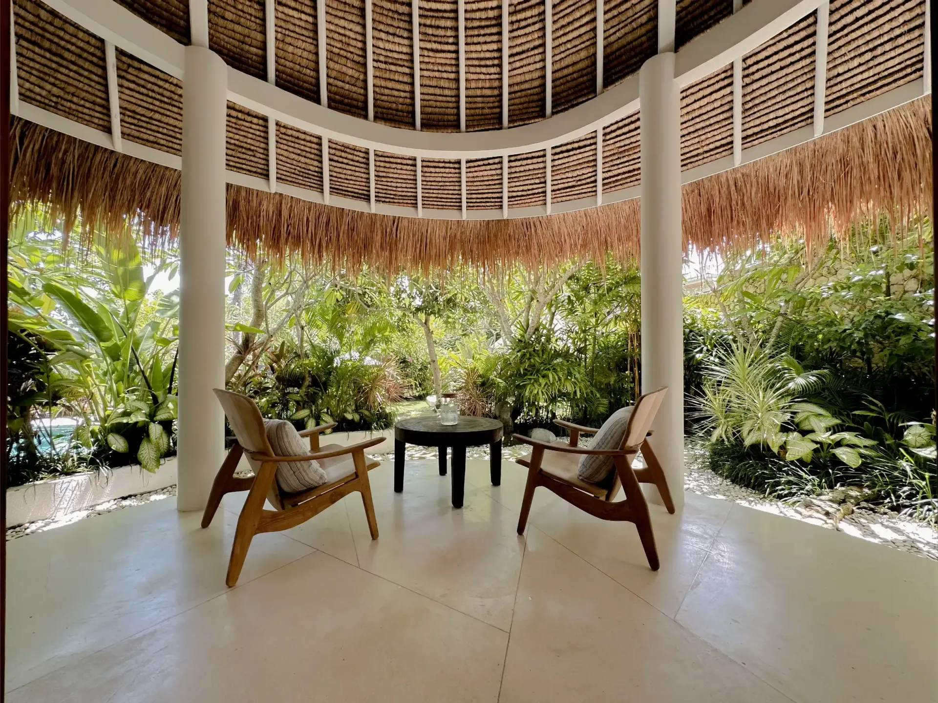 A covered outdoor seating area with a thatched roof, white pillars, and wooden chairs. Green foliage surrounds the space.