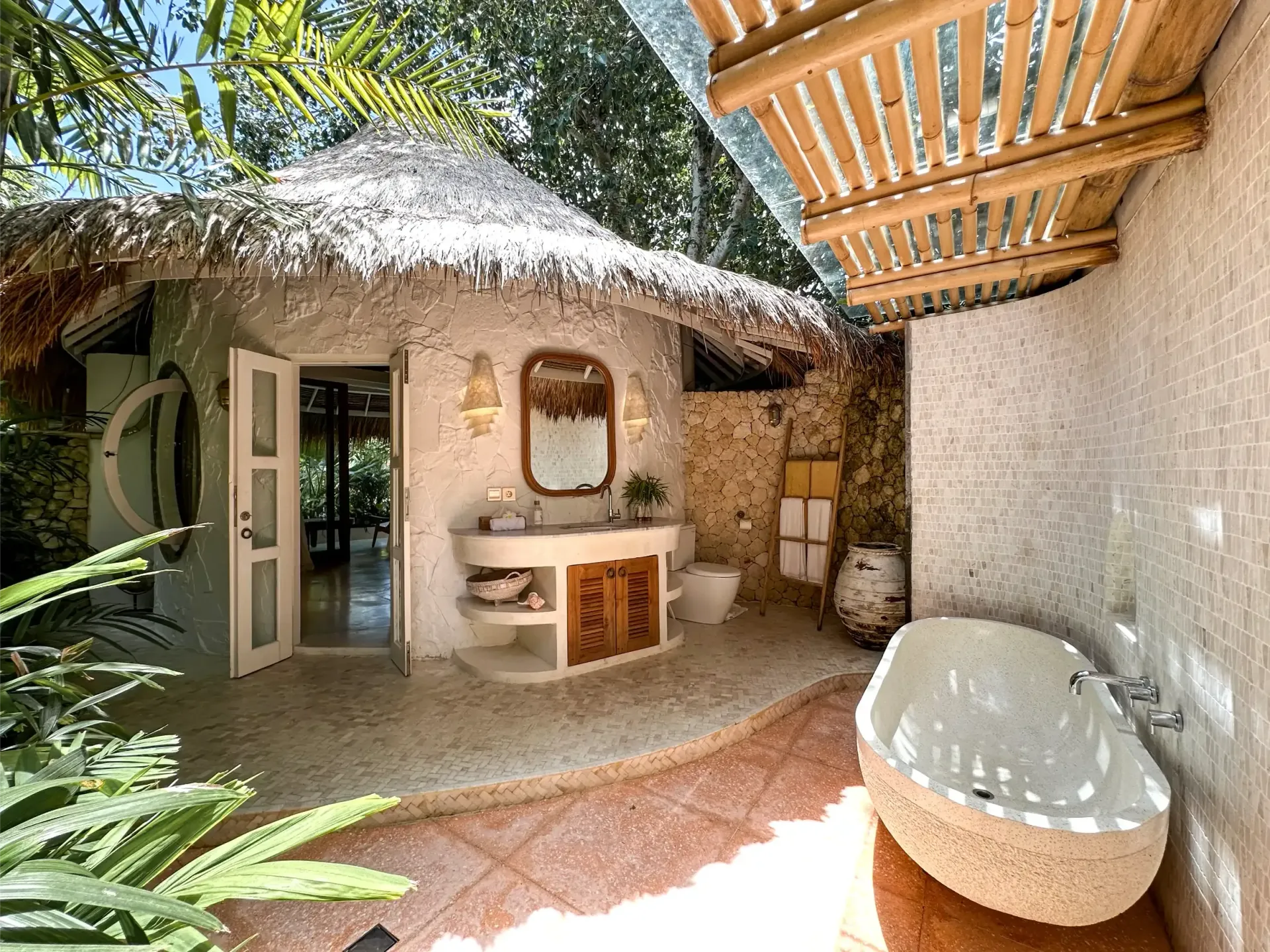 An outdoor bathroom with a thatched roof, stone walls, a bathtub, and a vanity. Tropical plants surround the space.