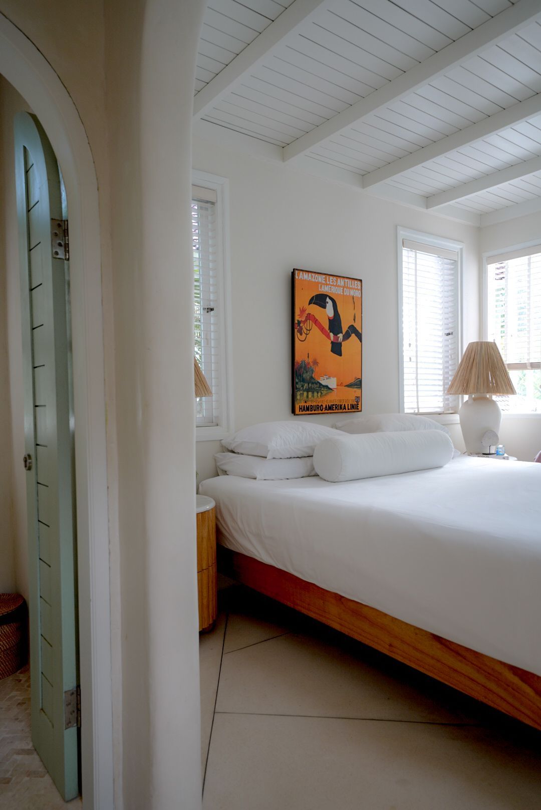 Bedroom interior with a white bed, toucan art, and arched doorway with a pale green door. 
