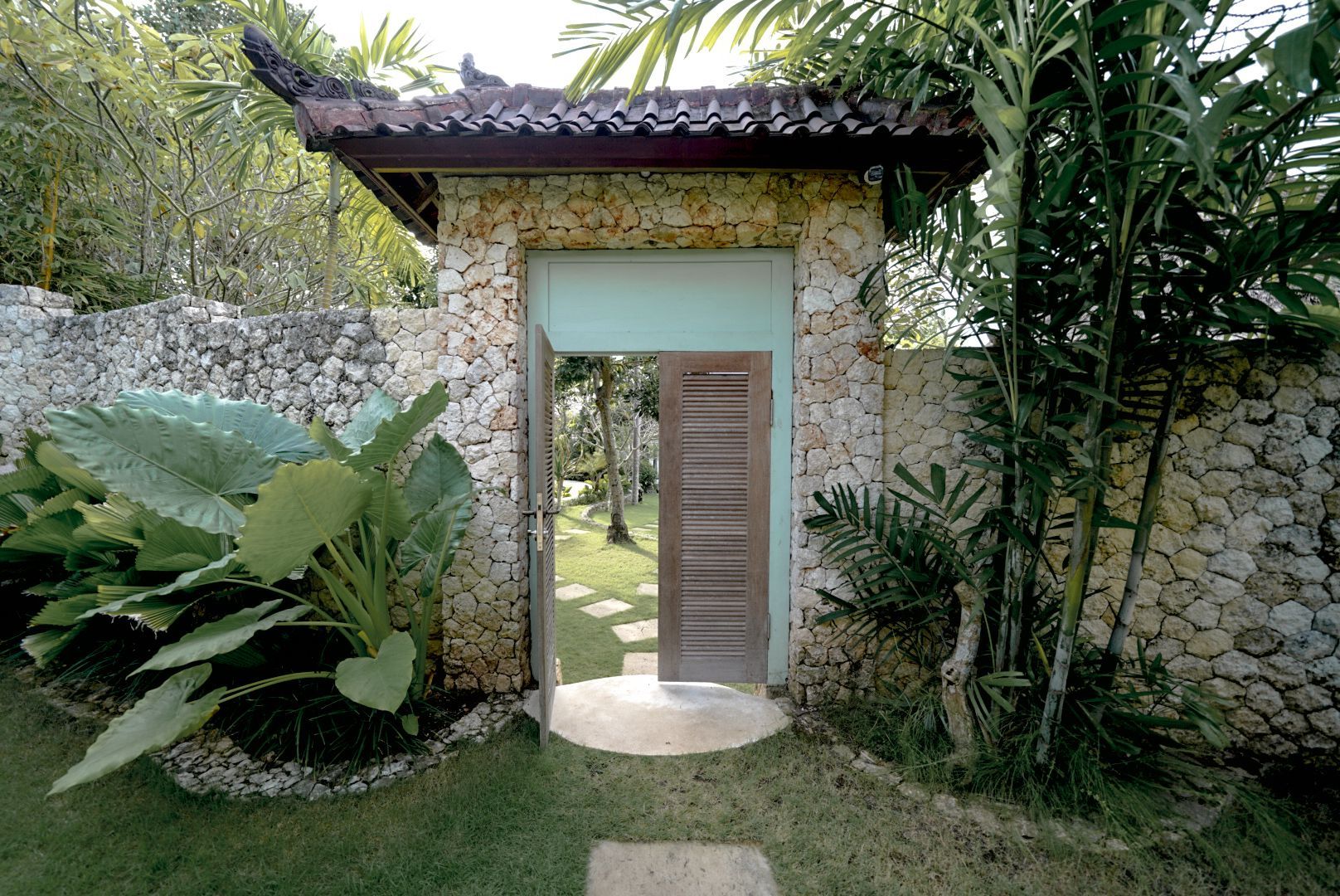 Stone archway entrance with a wooden door, leading to a grassy garden. Lush green plants surround the entrance.