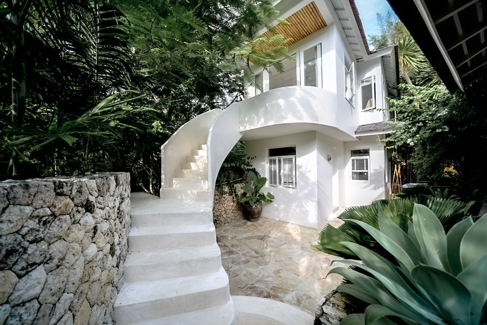 White house with curved staircase, surrounded by lush green foliage and stone wall.