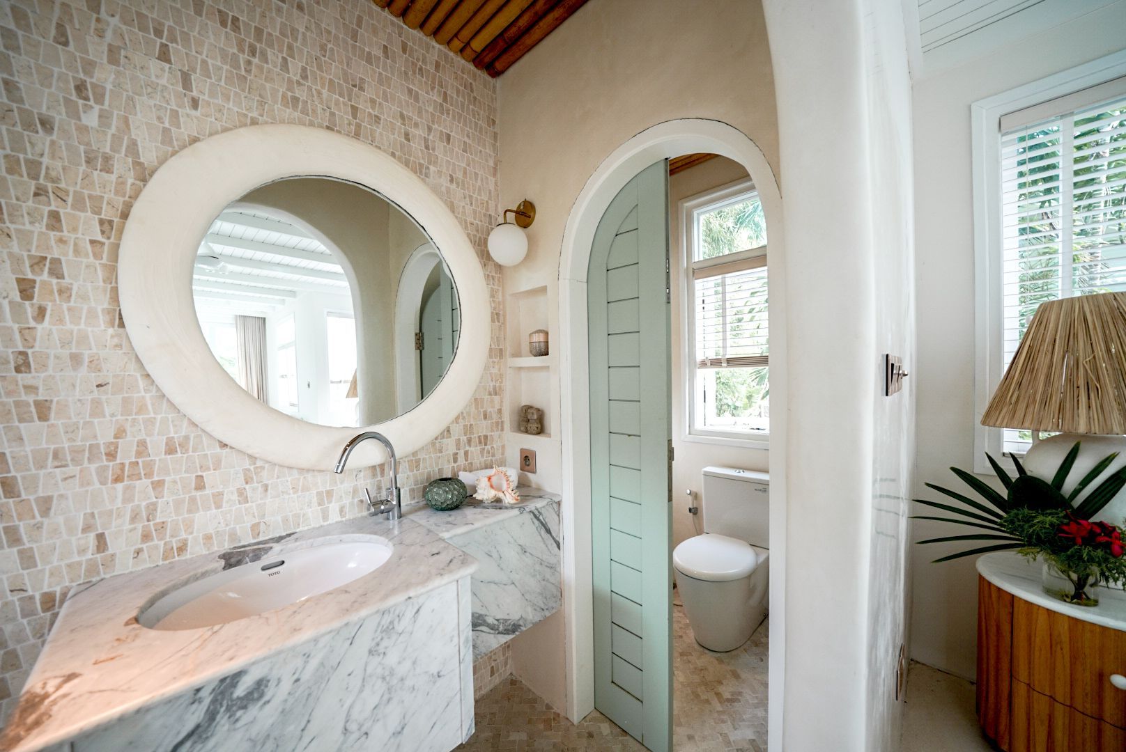 Bathroom with marble countertop, mosaic tile wall, arched doorway, and round mirror. Light and airy with natural light.