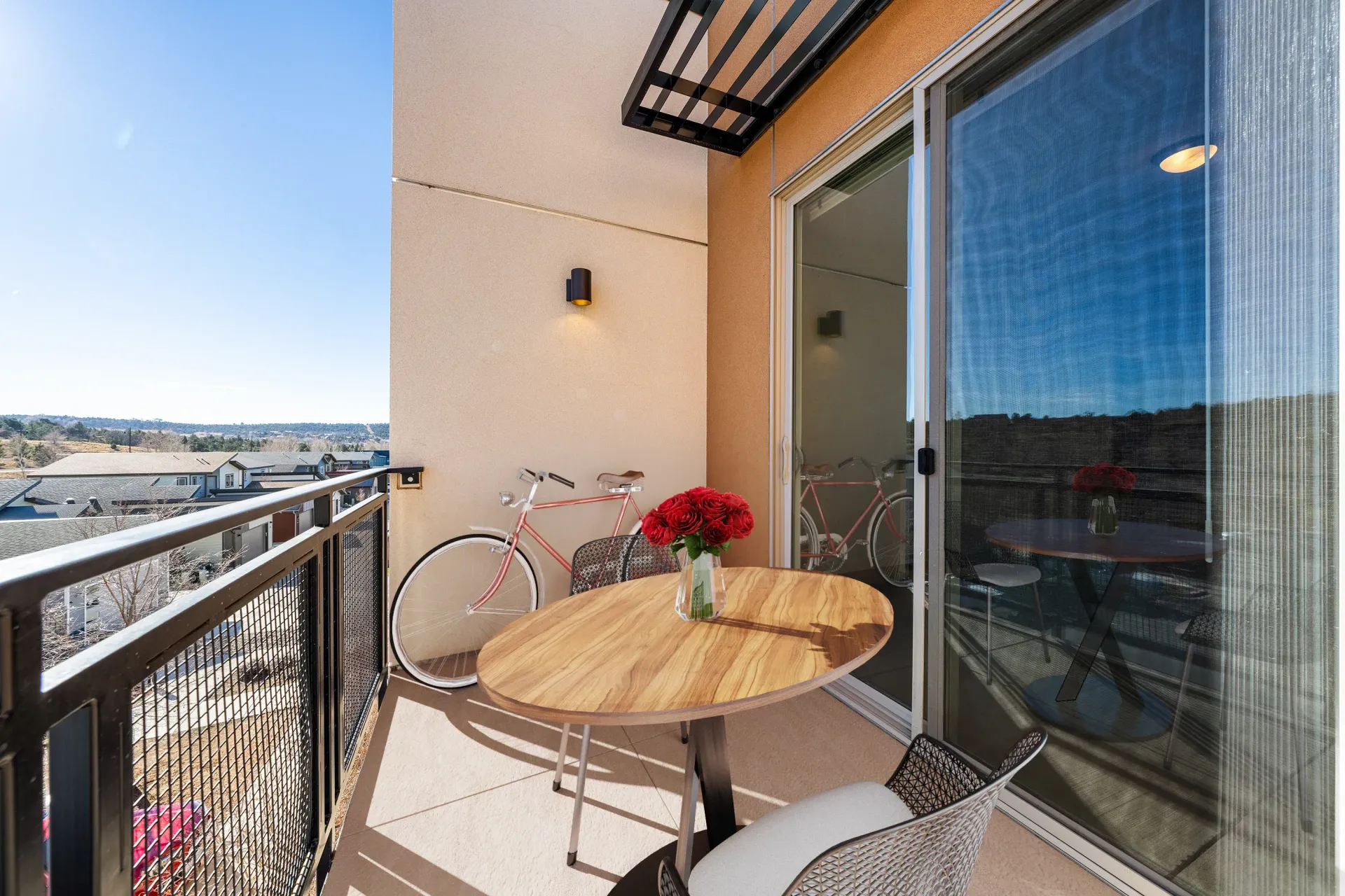 Balcony of an apartment with a round table, wicker chair, and a pink bicycle by the sliding glass door.