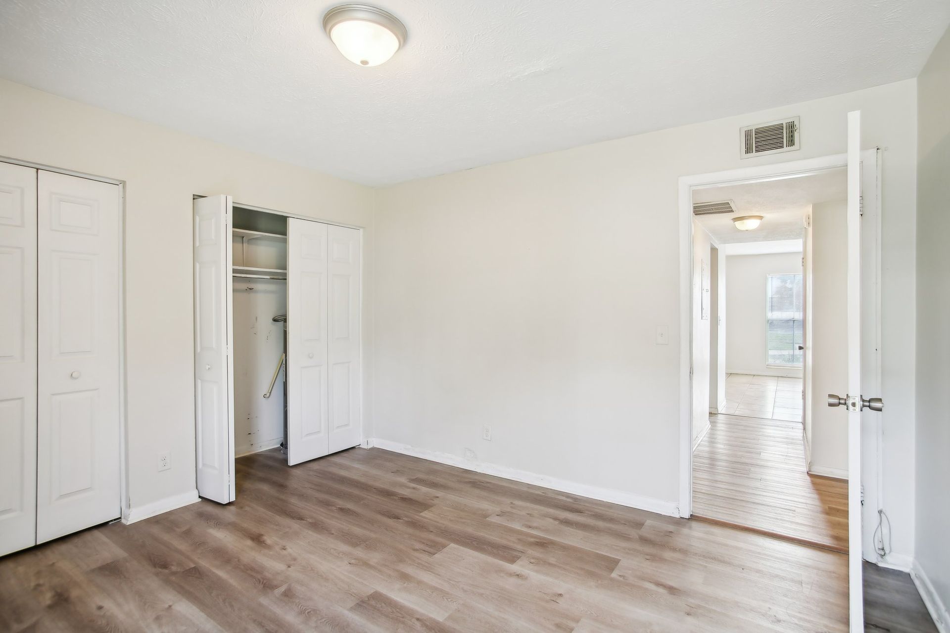 Empty bedroom with white walls, light wood-look flooring, and bi-fold closet doors. An open doorway leads to a hallway.