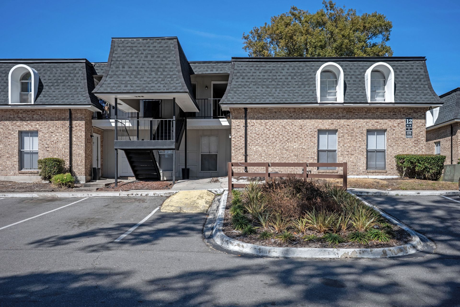 Apartment complex with brick exteriors and dark gray roofs; parking area in foreground, blue sky.