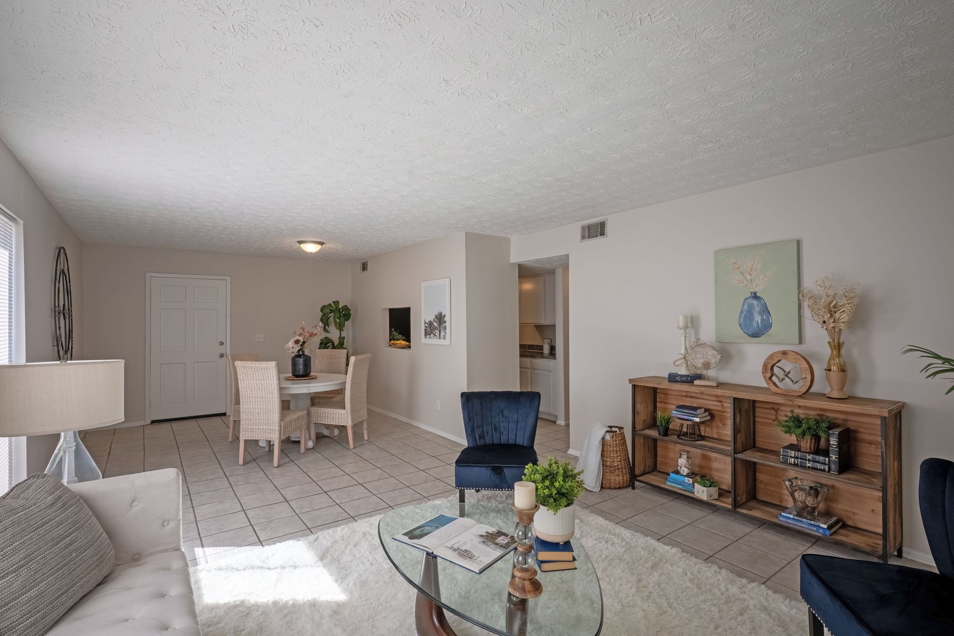 Living room with dining area, cream walls, white rug, blue accents, glass coffee table, and wood shelves.