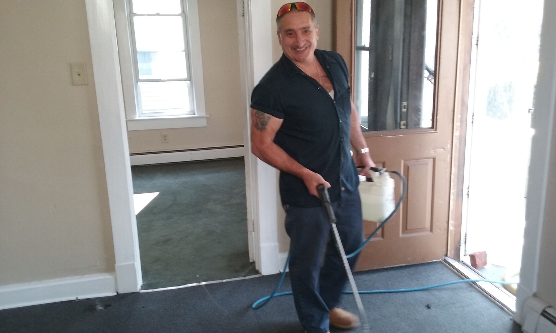A smiling person wearing safety glasses and a dark shirt holds a spray wand while cleaning a carpeted floor in a room.