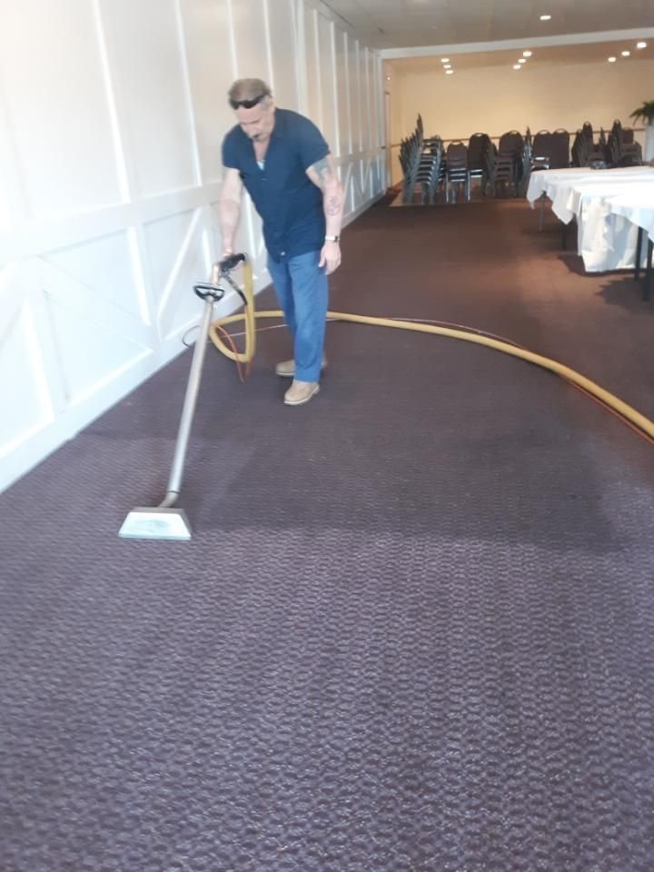 A person uses a commercial steam cleaning wand to clean dark patterned carpet in a banquet hall.