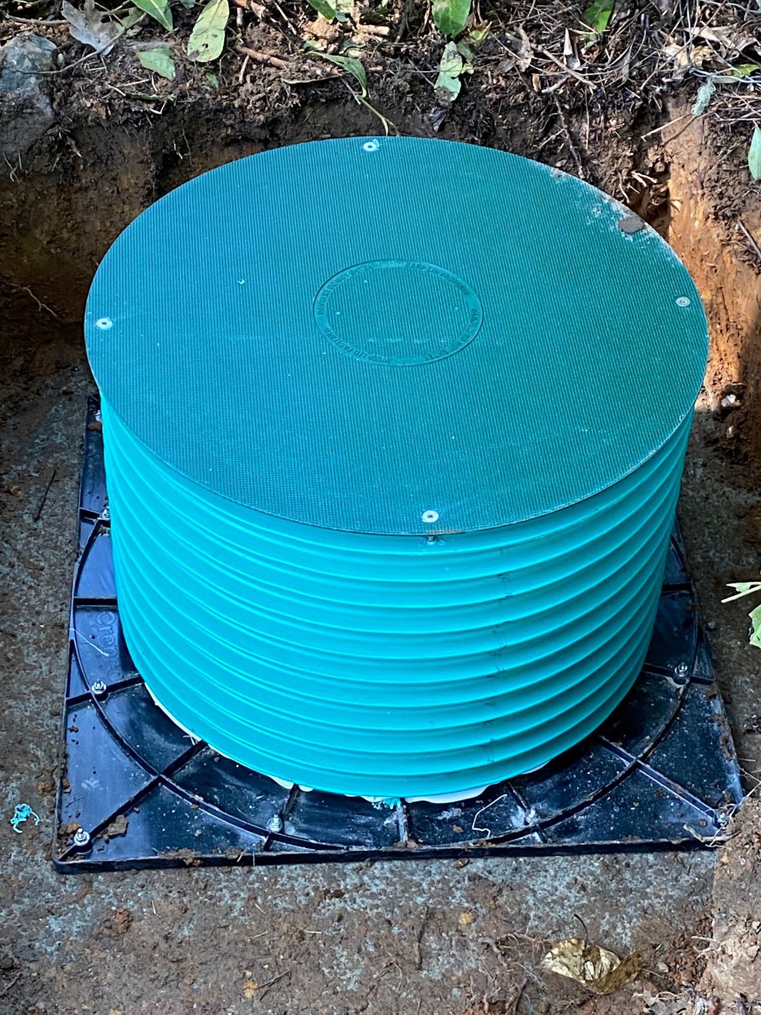 Green cylindrical container with a matching lid, sitting on a black square base in a dirt hole.