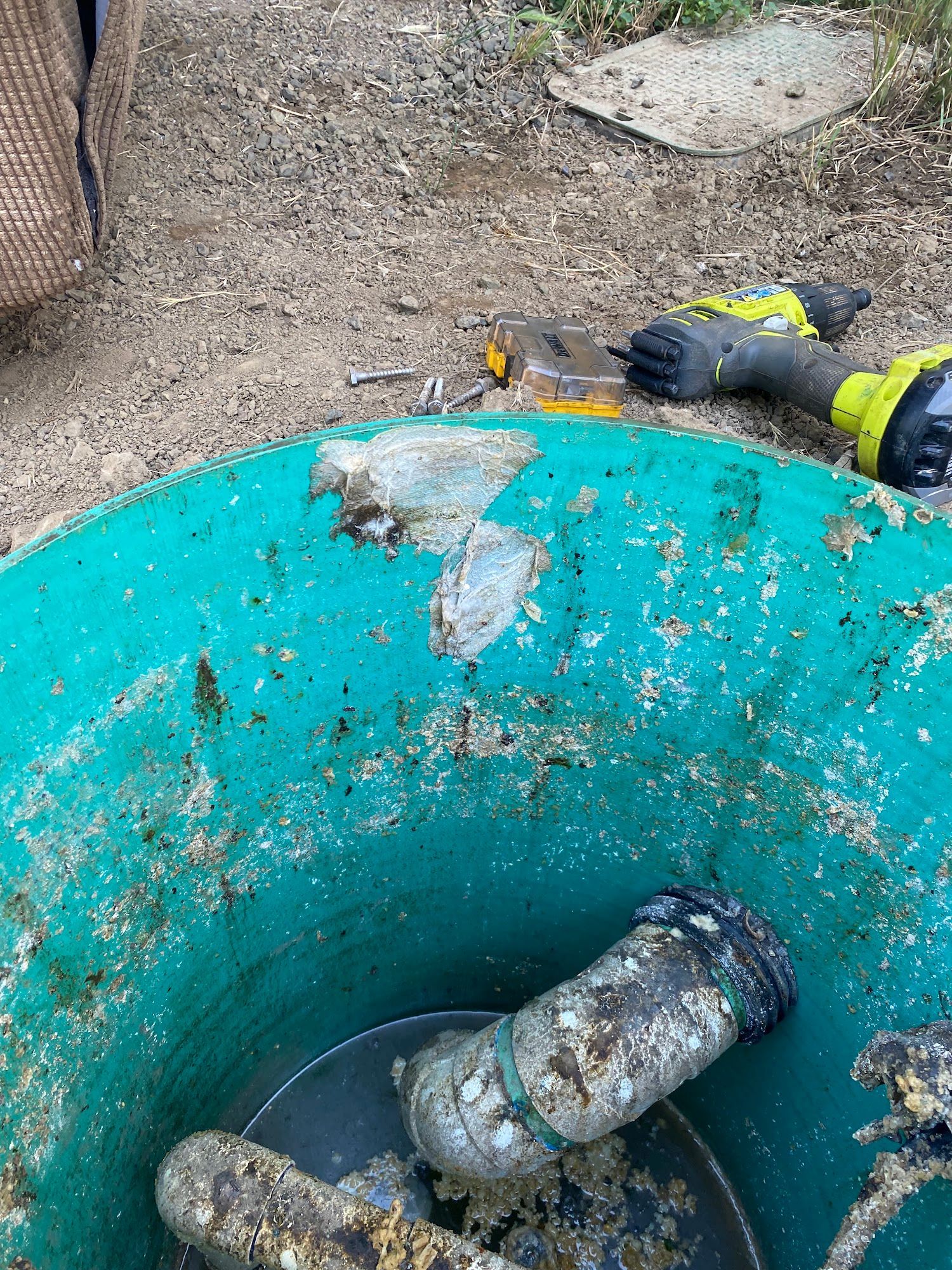 Green container with pipes, tools, and gloved hand on the rim, outdoors.