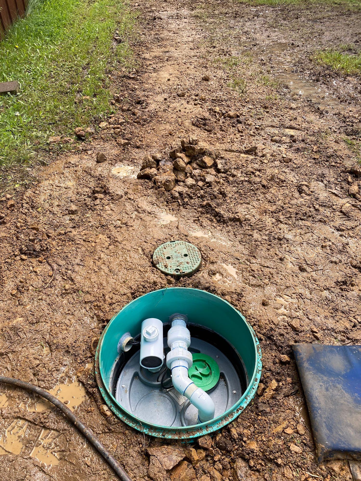 Open septic tank surrounded by dirt. Green lid off, visible pipes. Ground muddy.