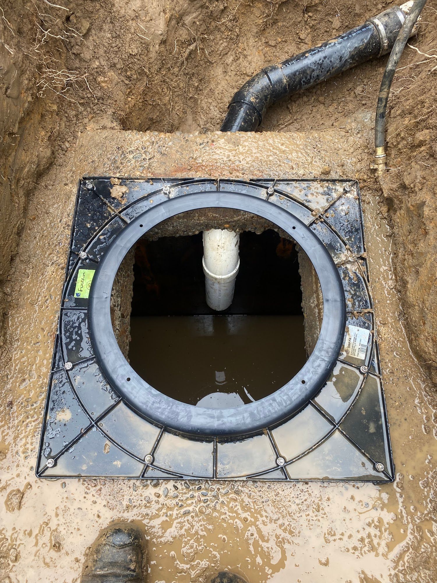 Open pit with black plastic frame and drain, white pipe, and water inside. Ground around is wet dirt.