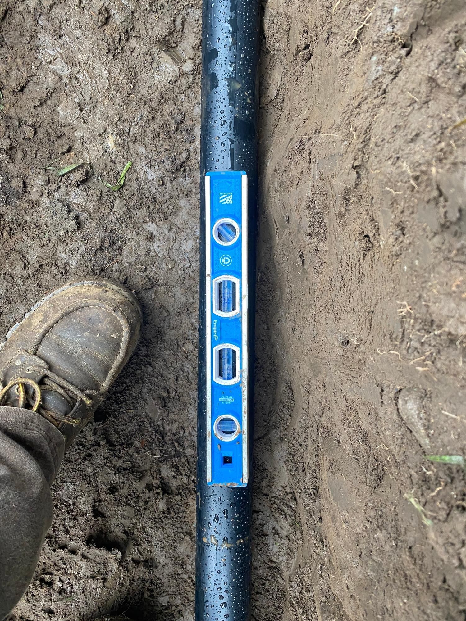 A black pipe in a trench, being checked for level with a blue level tool; foot visible.