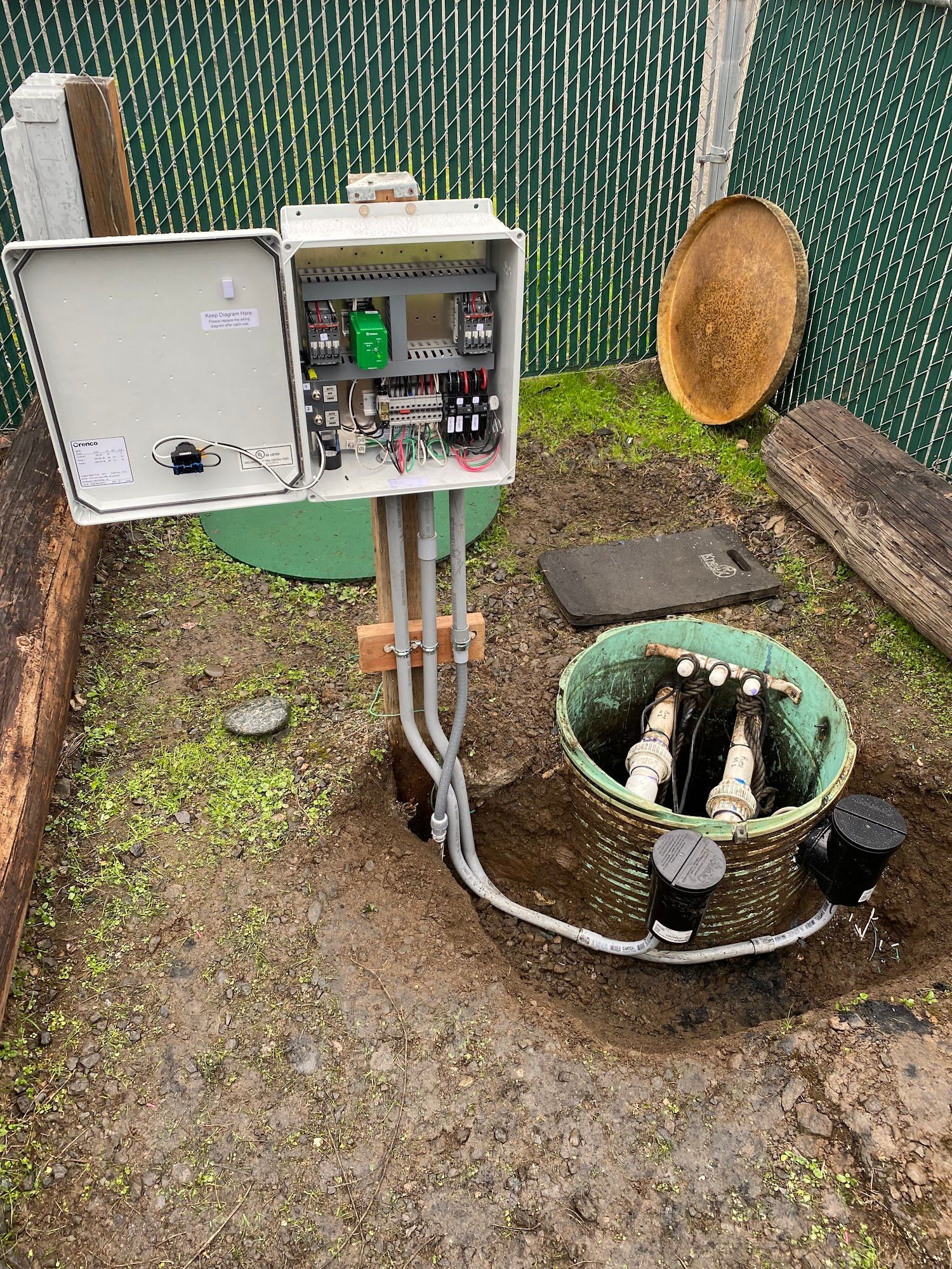 Control box with wires, open septic tank with pipes, and electrical equipment in a yard.