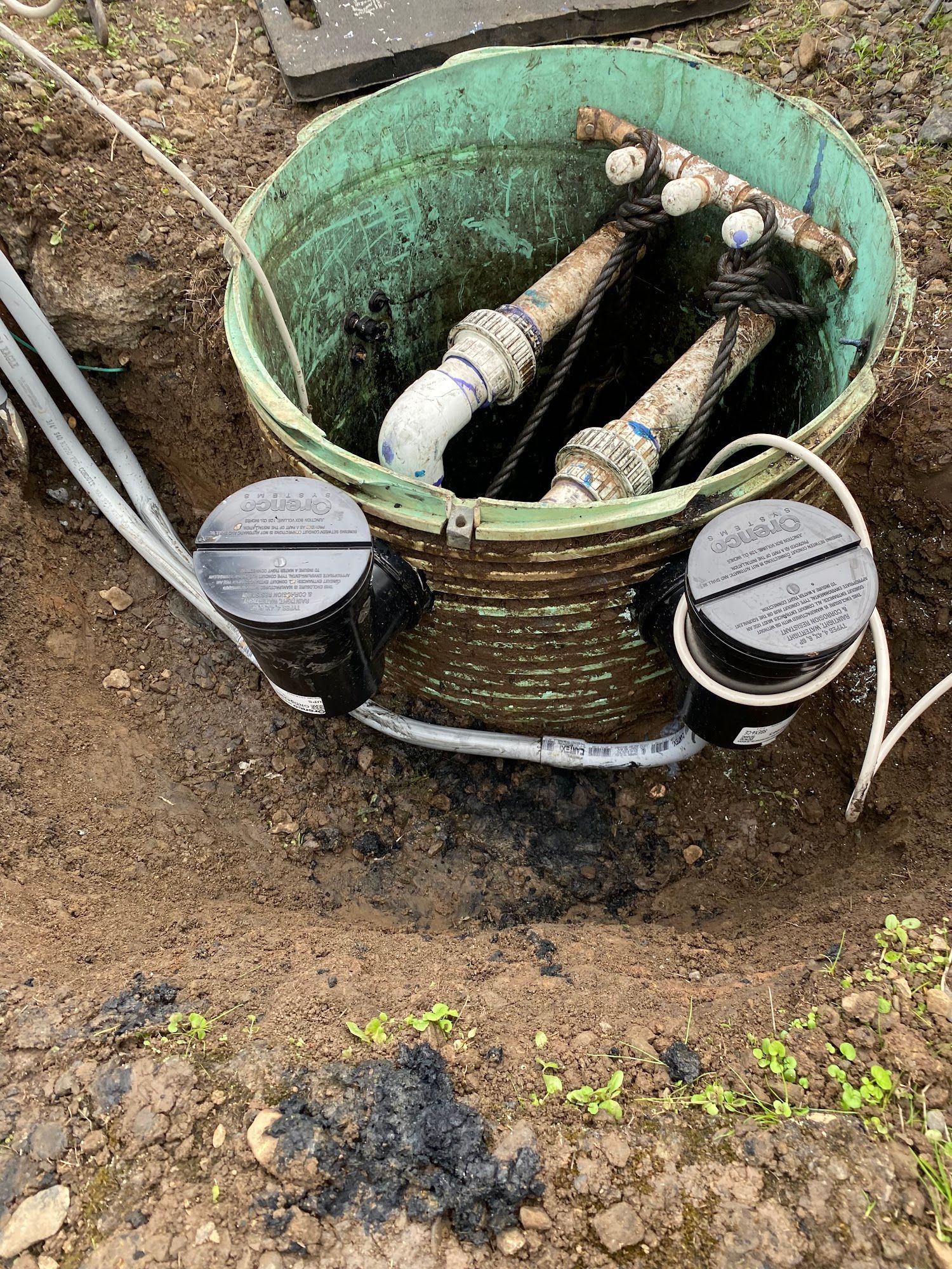 Open pit containing pipes and electrical components; a green bucket sits in the pit.