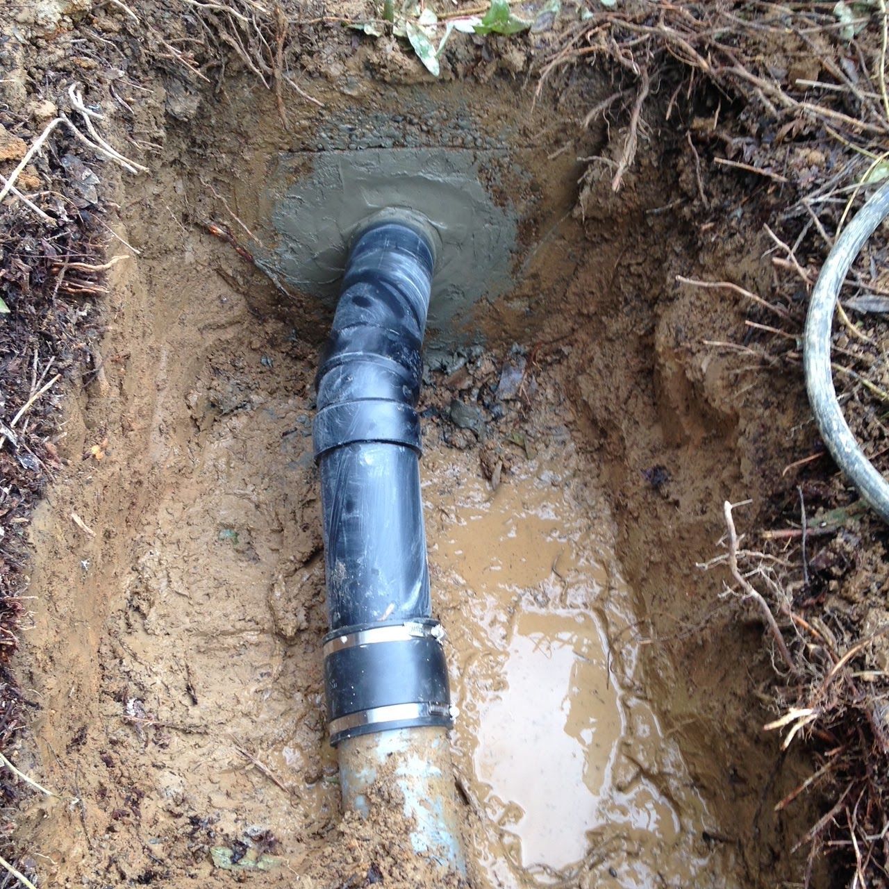 Black pipe in an excavated hole, connected with clamps, surrounded by soil and mud.