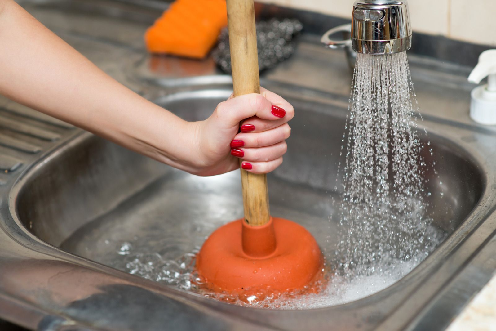 Hand holding a plunger under running water in a stainless steel sink