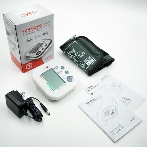 Kit de monitor de presión arterial con brazalete, adaptador y manual de instrucciones sobre un fondo blanco.