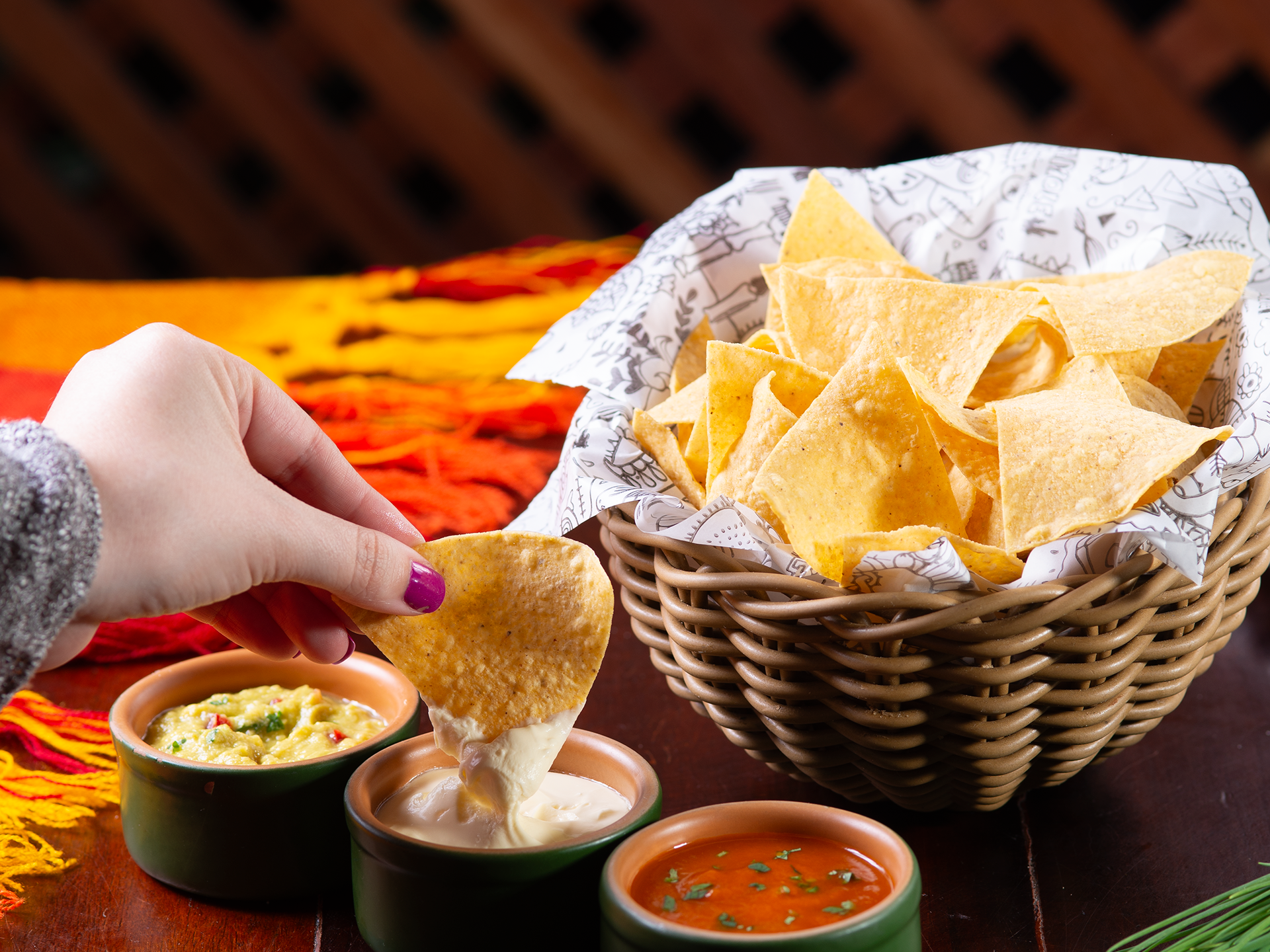 Uma mão mergulha um nacho em uma tigela de molho cremoso ao lado de salsa e uma cesta de nachos.