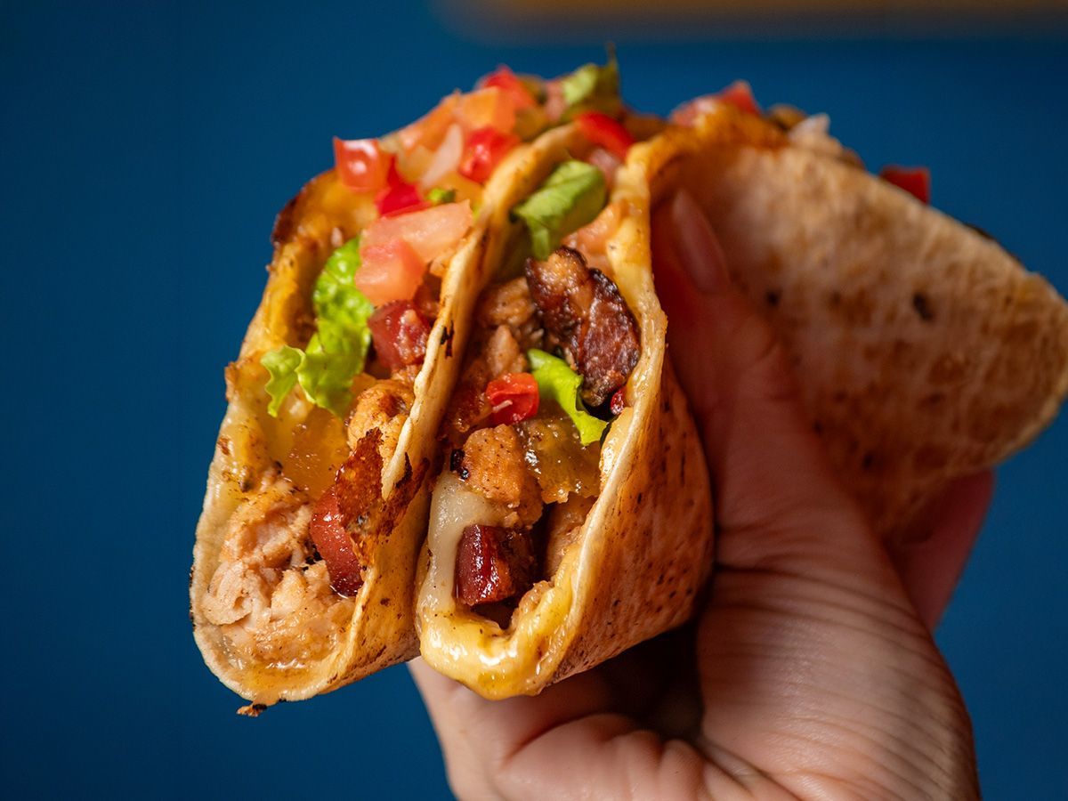 Mão segurando um taco recheado com carne, alface e tomate contra um fundo azul.