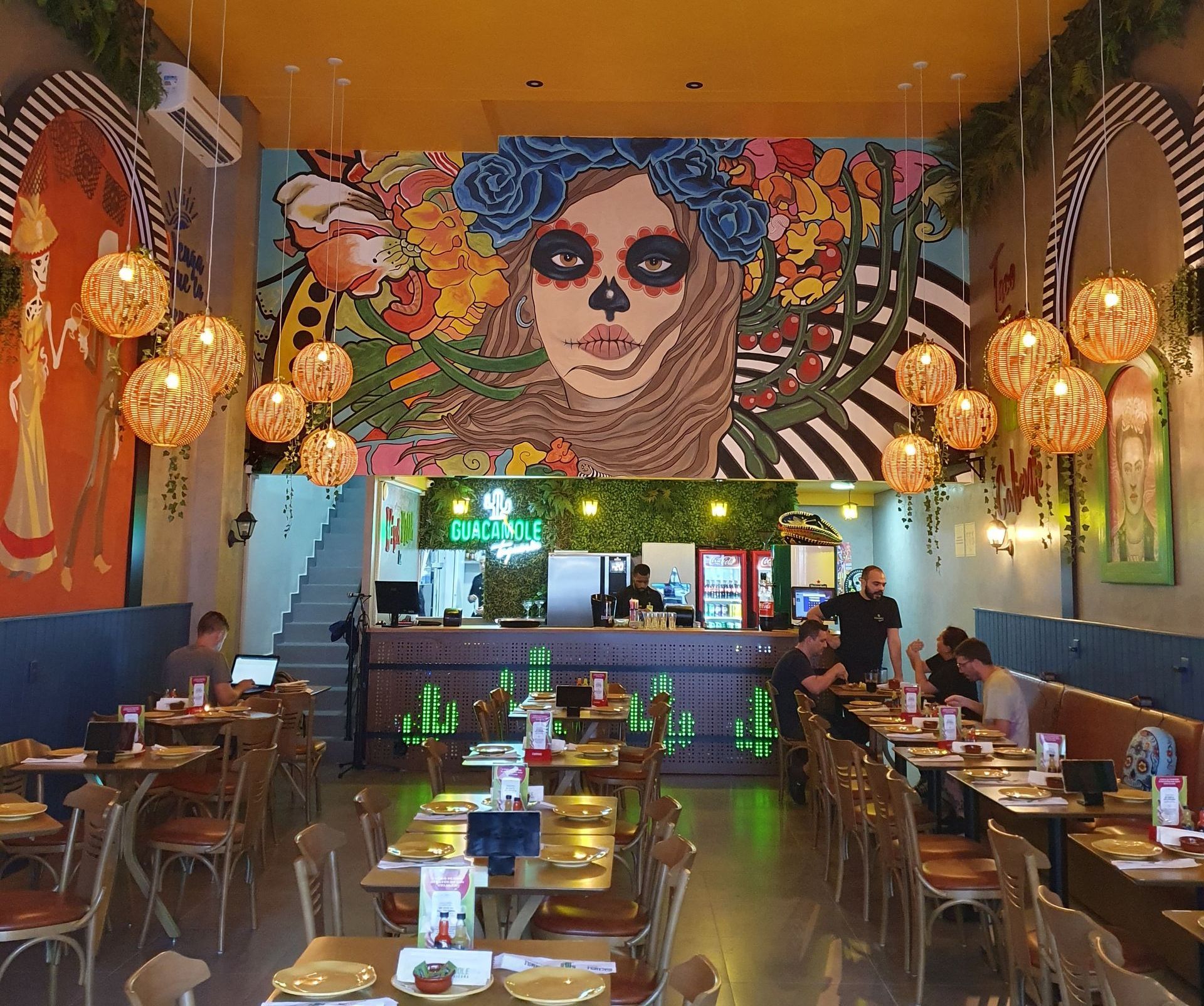 Interior colorido de restaurante com um grande mural, lanternas penduradas e clientes sentados às mesas.