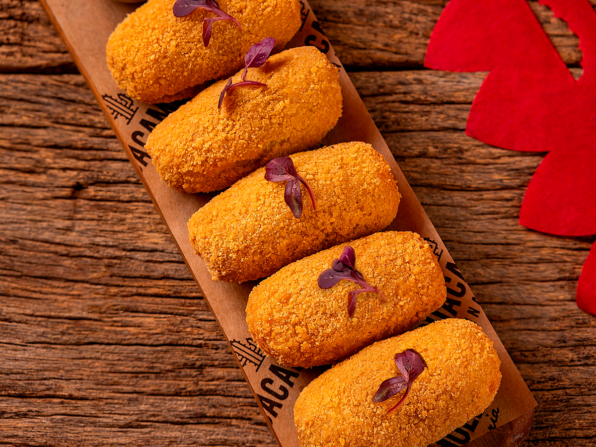 Croquetes dourados empanados em uma bandeja de madeira com cobertura vermelha sobre uma mesa rústica de madeira.