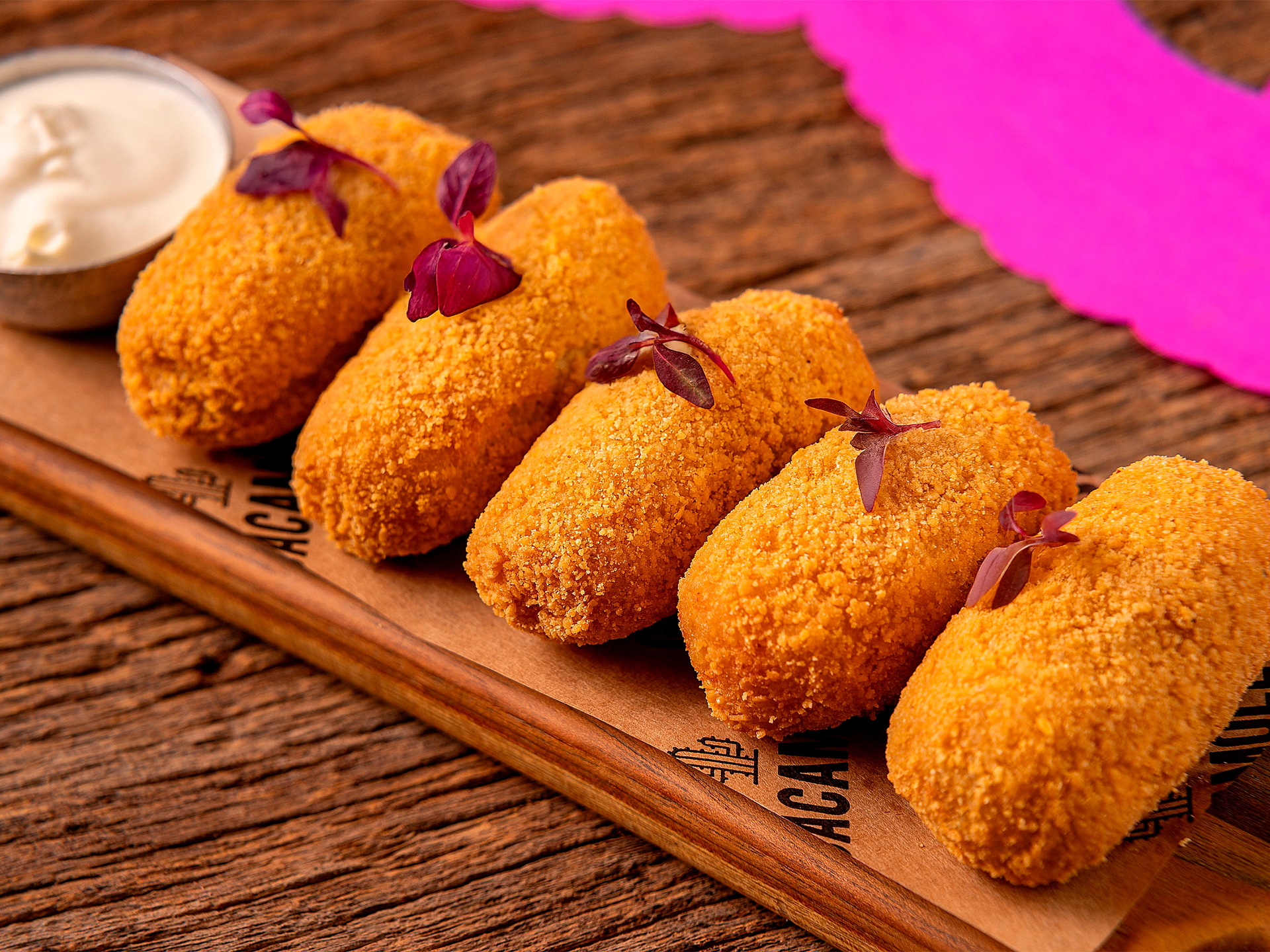 Croquetes dourados empanados em uma tábua de madeira com molho para mergulhar, em uma mesa rústica.