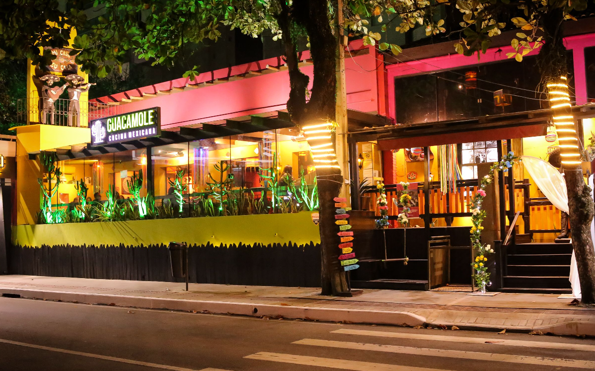 Pátio de restaurante colorido e iluminado à noite, com mesas ao ar livre e toldo rosa em uma rua da cidade.