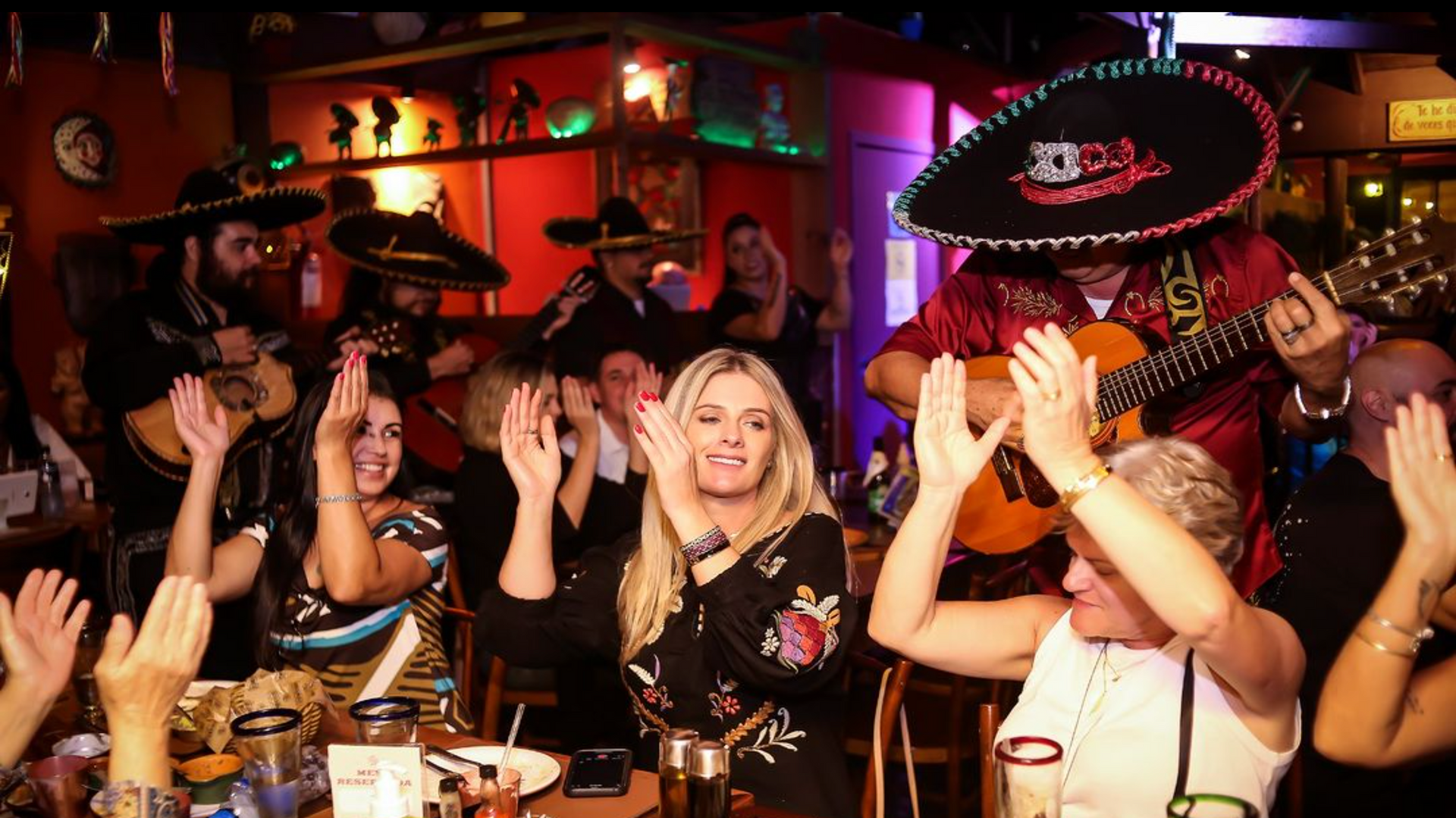 Pessoas vibrando em um bar animado, com um músico mariachi de sombrero ao lado delas.