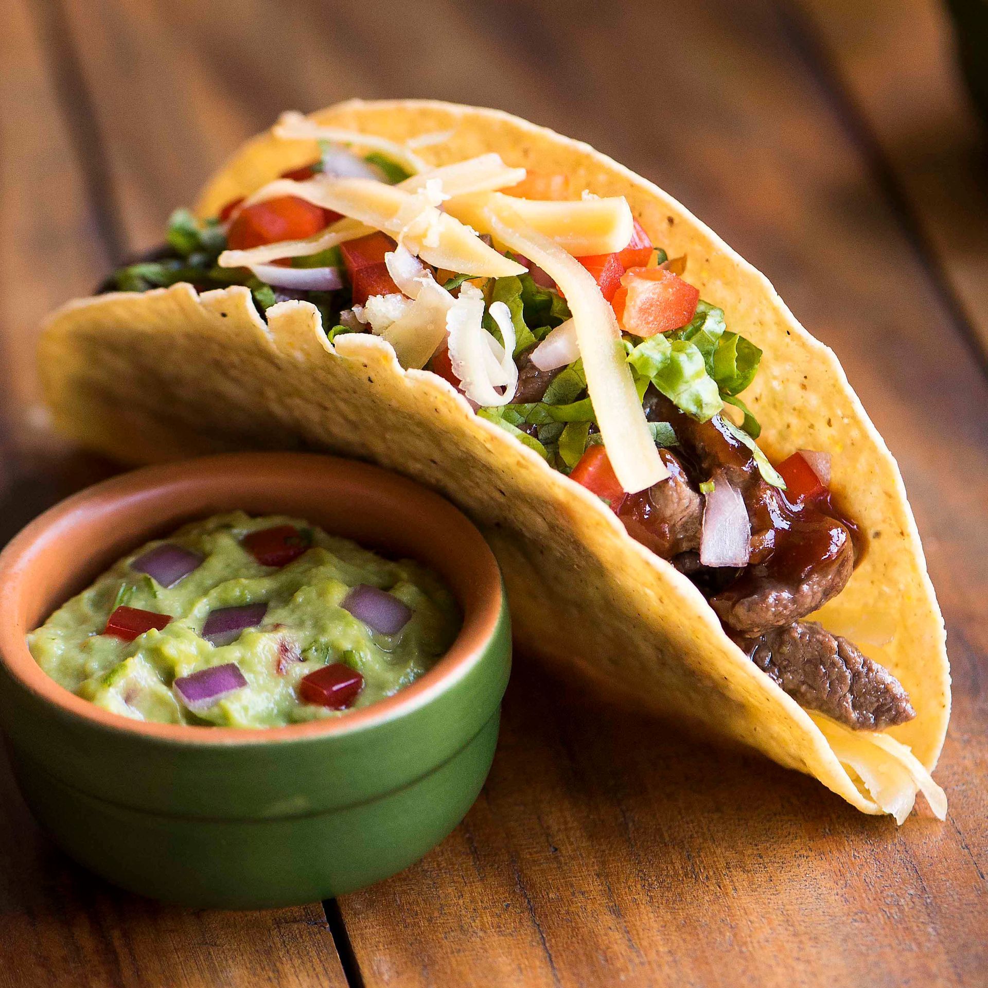 Taco de carne com alface, tomate e queijo ao lado de uma tigela de guacamole em uma mesa de madeira.