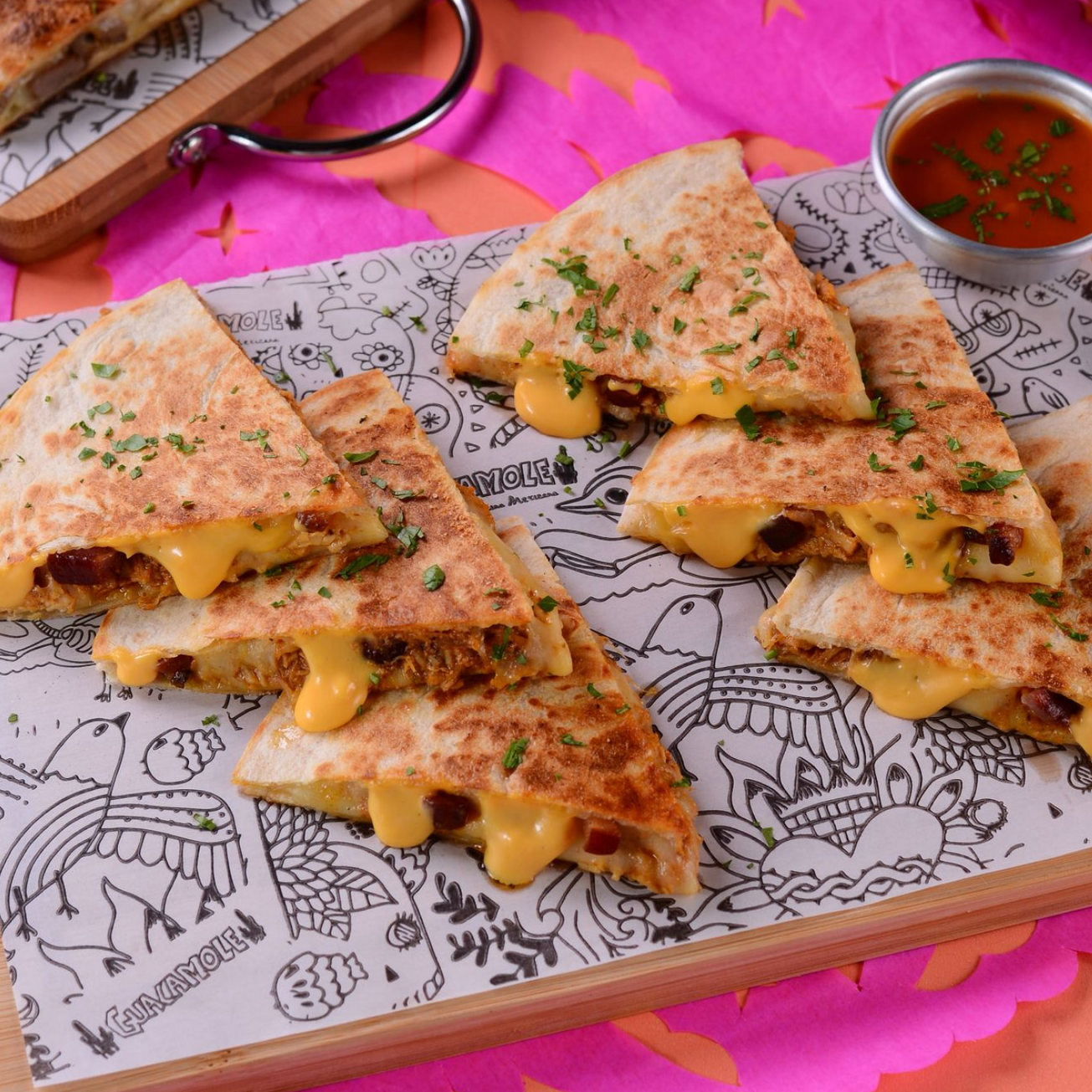 Fatias de quesadilla tostadas e recheadas com queijo, servidas em uma bandeja estampada com salsa, sobre um fundo rosa vibrante.