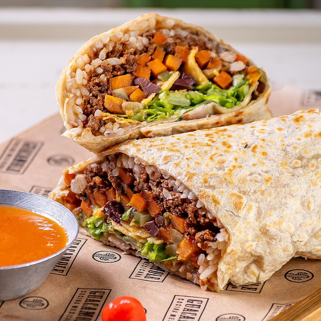 Burrito cortado com carne, alface e pimentões picados ao lado de um copo de molho vermelho em um wrap de papel.