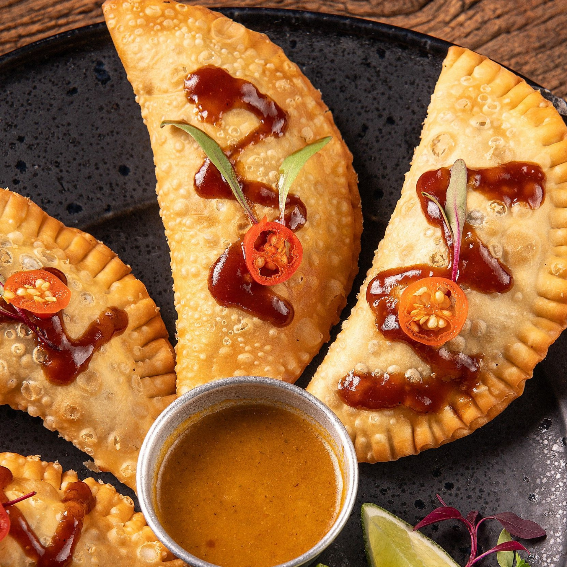 Três pastéis fritos com chutney e pimenta servidos em um prato preto, com molho para mergulhar no centro.