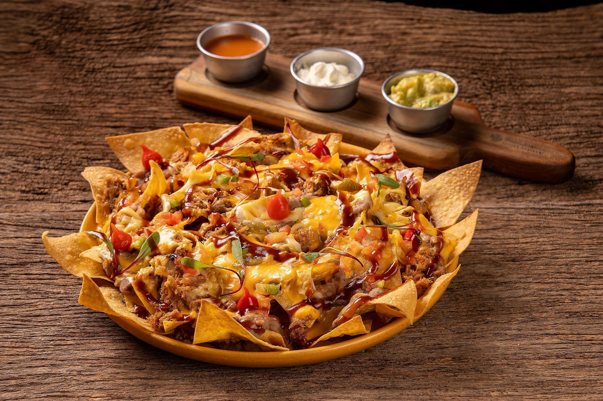 Nachos recheados com queijo, tomates e pimentões, servidos em uma mesa rústica com três molhos para mergulhar.