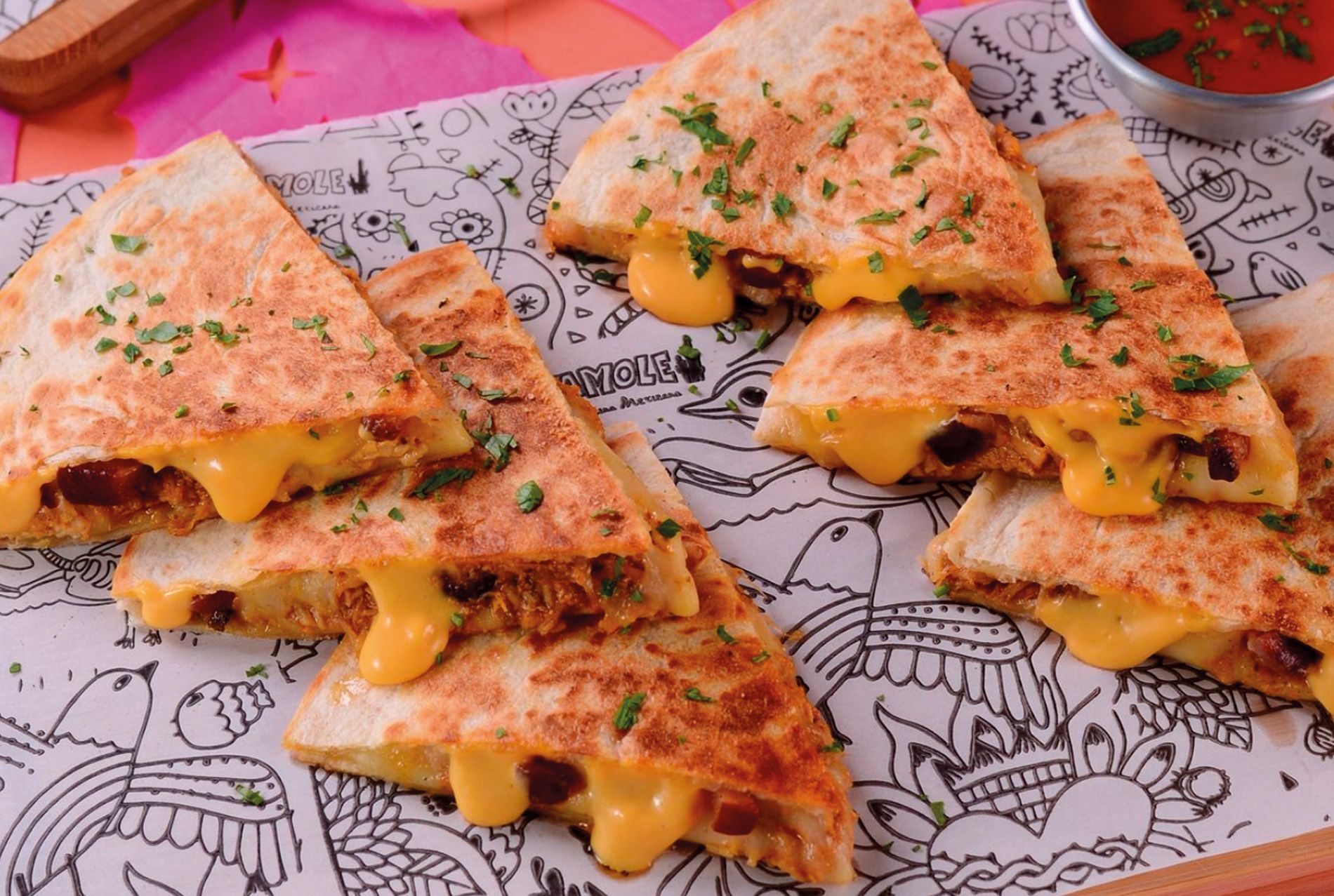 Três fatias de quesadilla com queijo, servidas em papel estampado, acompanhadas de um pequeno copo de molho vermelho.