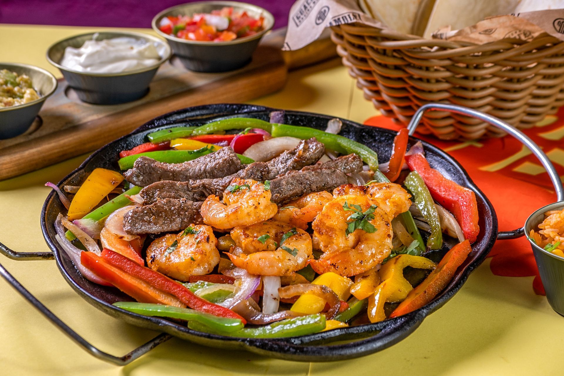 Uma travessa colorida com carnes e legumes grelhados sobre uma mesa, acompanhada de guarnições e uma cesta de vime por perto.