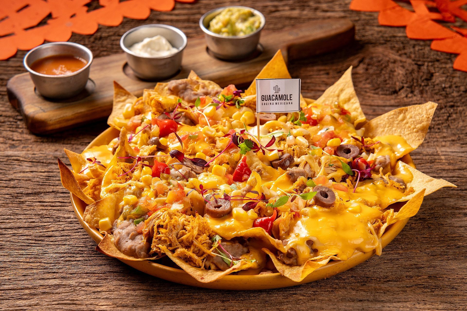 Nachos recheados com queijo derretido, acompanhamentos e três molhos para mergulhar, servidos sobre uma mesa de madeira.