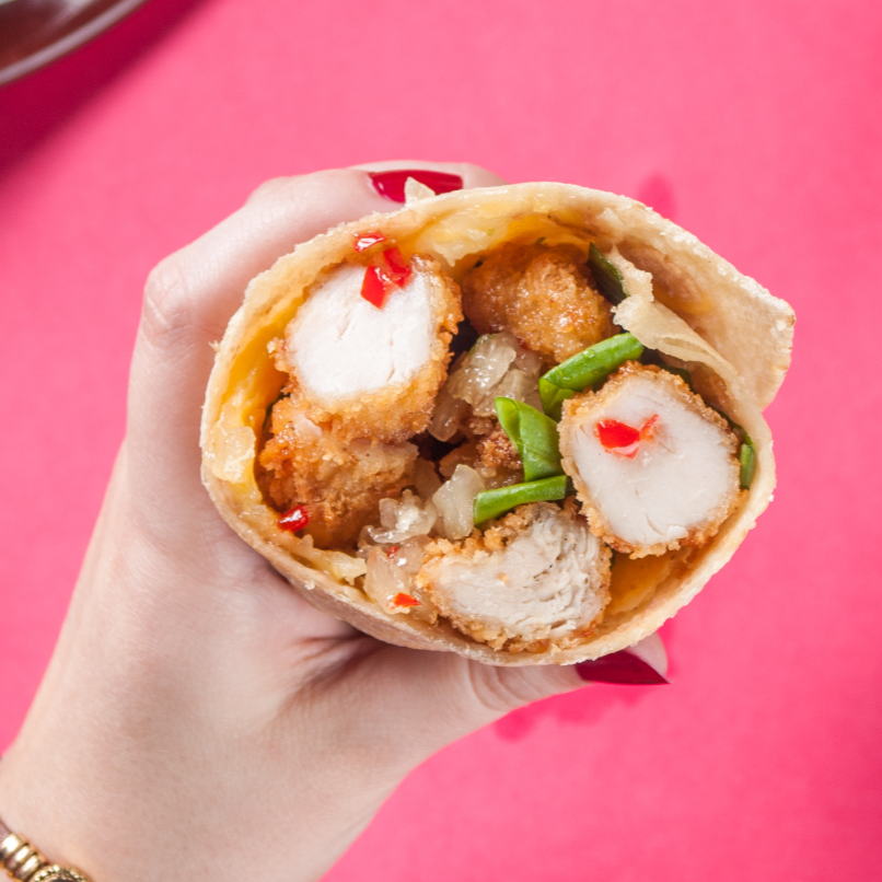 Mão segurando um wrap recheado com frango, arroz, vagem e molho vermelho contra um fundo rosa.