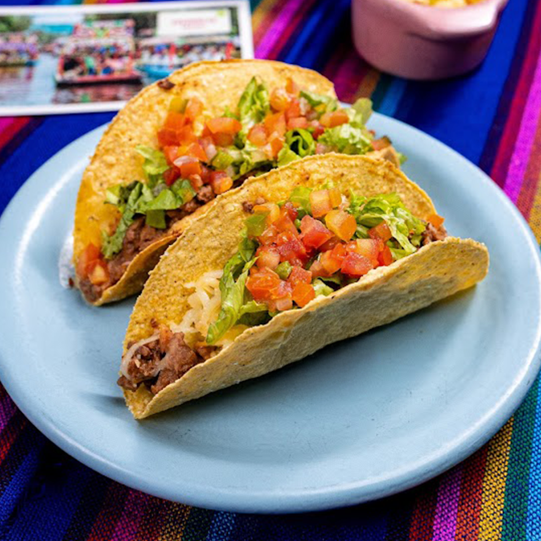Dois tacos de carne com alface e pico de gallo em um prato azul, sobre uma toalha de mesa listrada e colorida.
