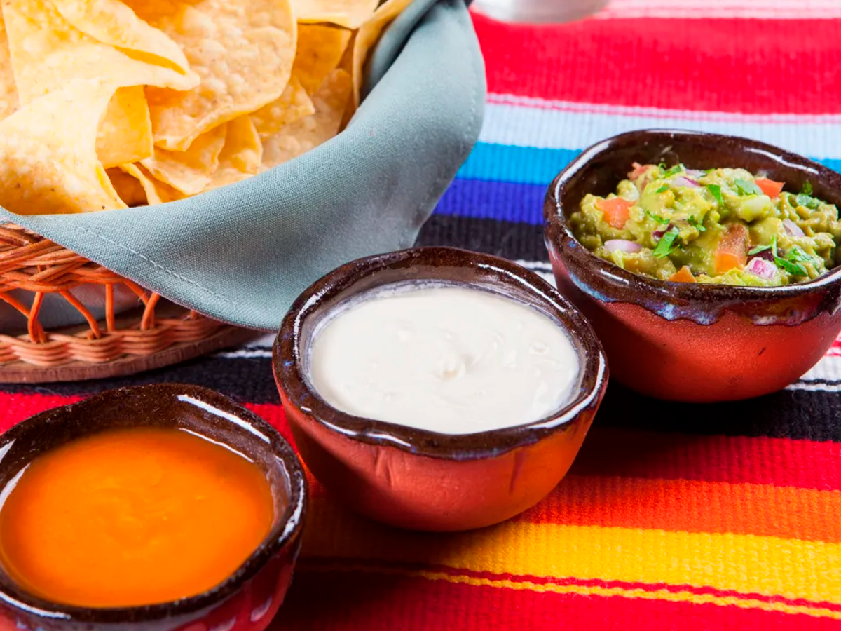 Três tigelas de salsa e molhos ao lado de nachos sobre um pano listrado colorido.