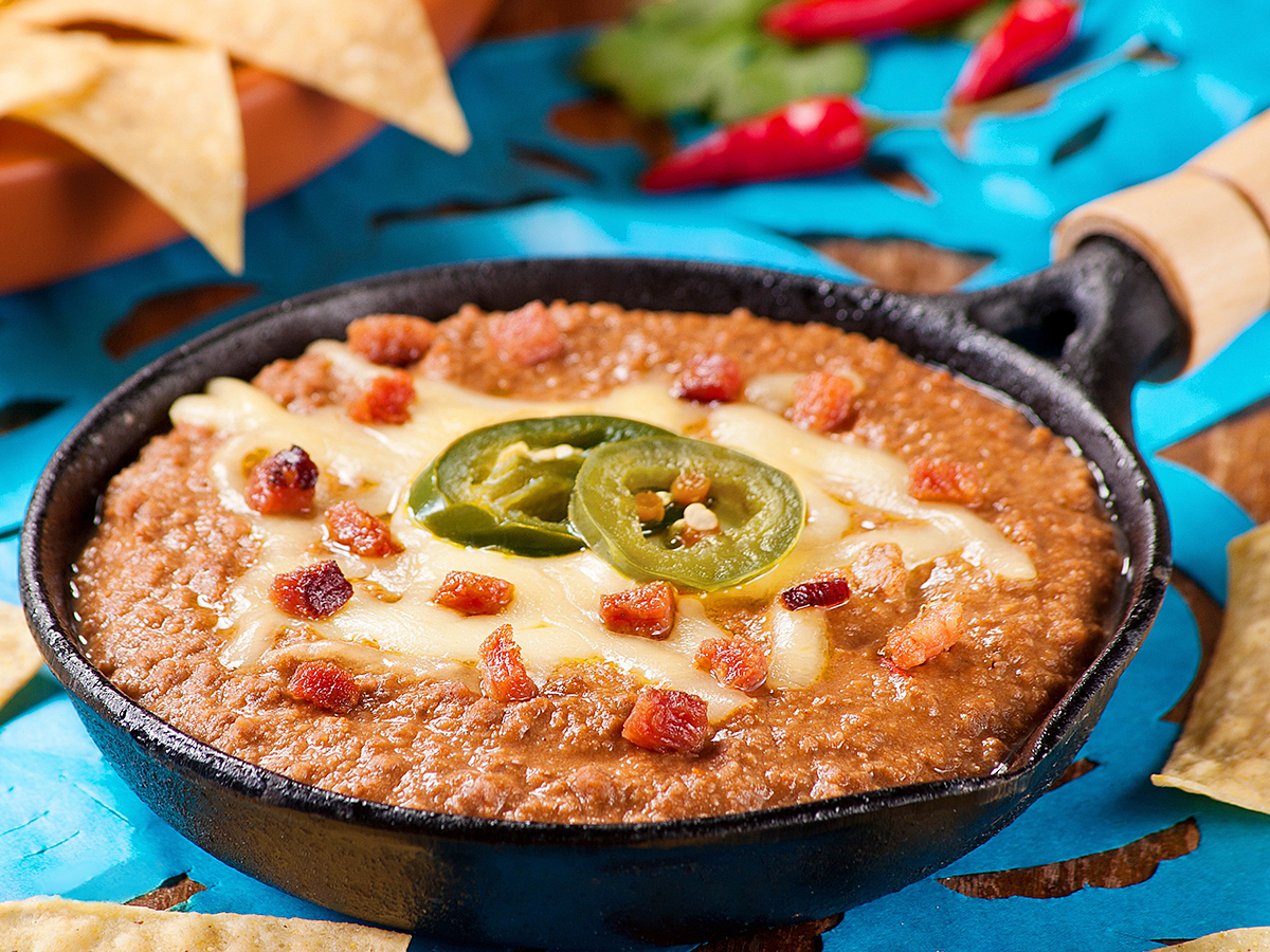 Frigideira com molho cremoso de feijão, coberta com fatias de jalapeño e pedaços de bacon sobre uma superfície azul.