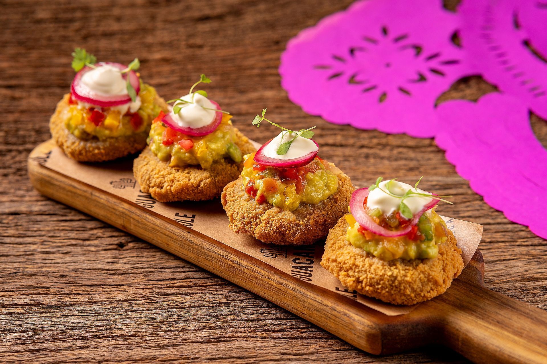 Quatro aperitivos fritos cobertos com fatias de rabanete, dispostos em uma tábua de madeira, com um guardanapo rosa sobre uma mesa rústica.