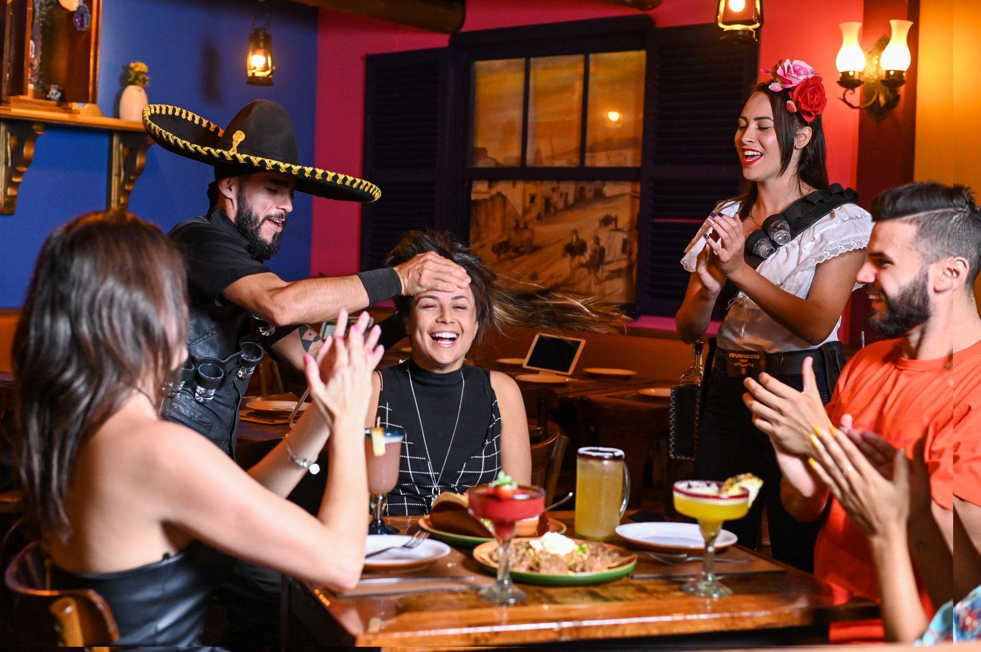 Amigos rindo e batendo palmas em uma mesa de jantar festiva em um restaurante aconchegante, com bebidas e um sombrero.
