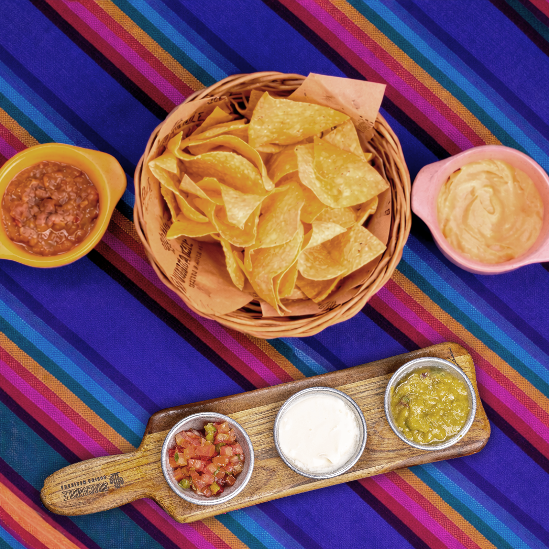 Três molhos e uma cesta de nachos sobre uma toalha listrada colorida.