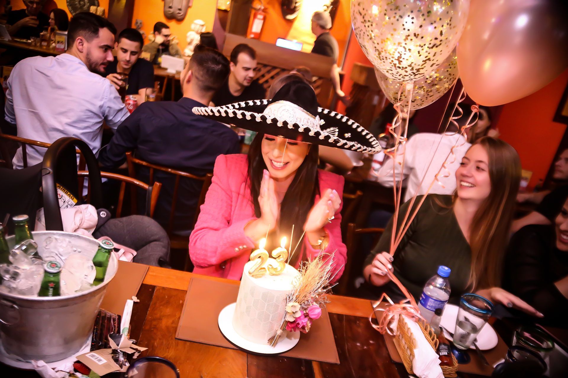 Comemoração de aniversário em um restaurante, com uma mulher de sombrero sorrindo ao lado de um bolo iluminado e balões.