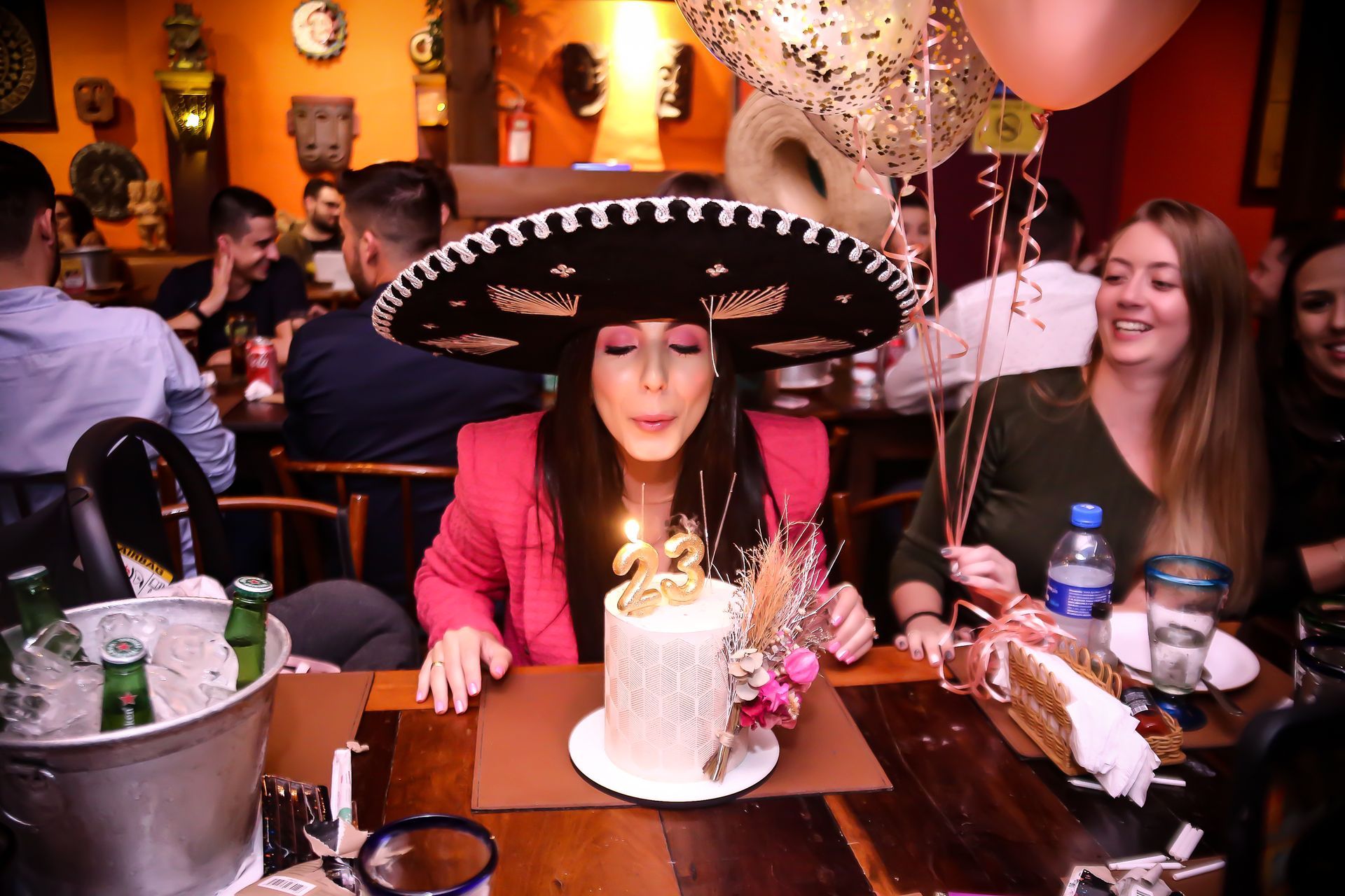 Mulher de jaqueta rosa e sombrero comemorando aniversário com um bolo em um restaurante animado.