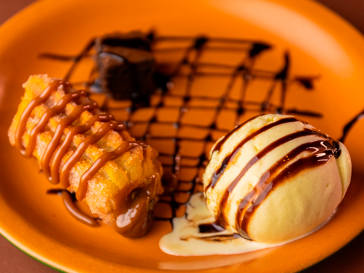 Duas sobremesas com cobertura de chocolate sobre um prato laranja com fundo quadriculado escuro.