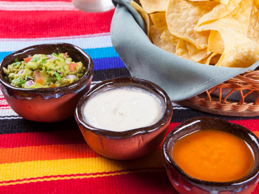 Três tigelas de salsa e molho para mergulhar, ao lado de nachos, sobre uma toalha de mesa listrada e colorida.